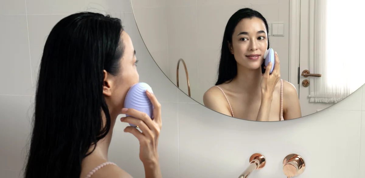 Woman at a bathroom mirror using a lavender FOREO LUNA device on her cheek