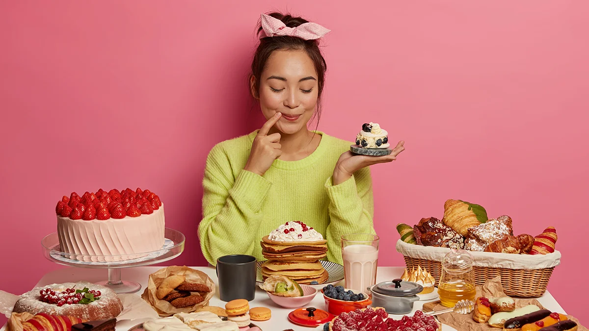 An Asian woman in yellow in front of a pink wall sitting at a full table of sweets