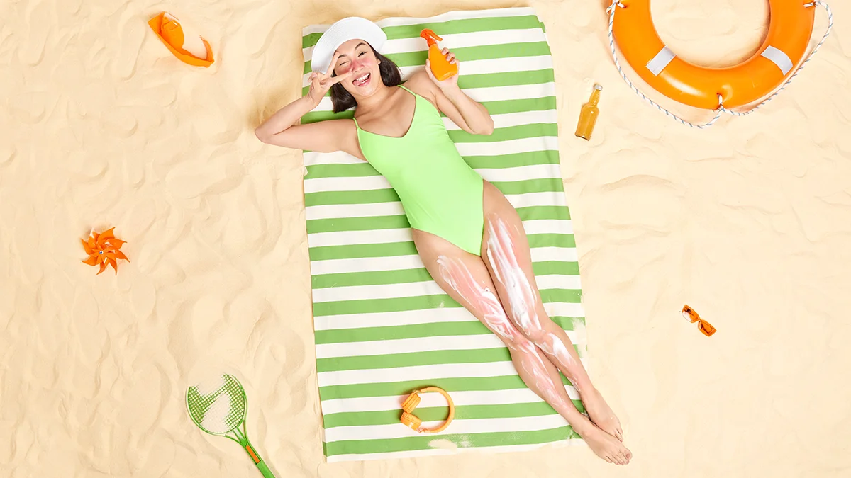 A young, beutiful Asian woman on a sandy beach, lying on a towel, birds perspective, with SPF cream all over her legs
