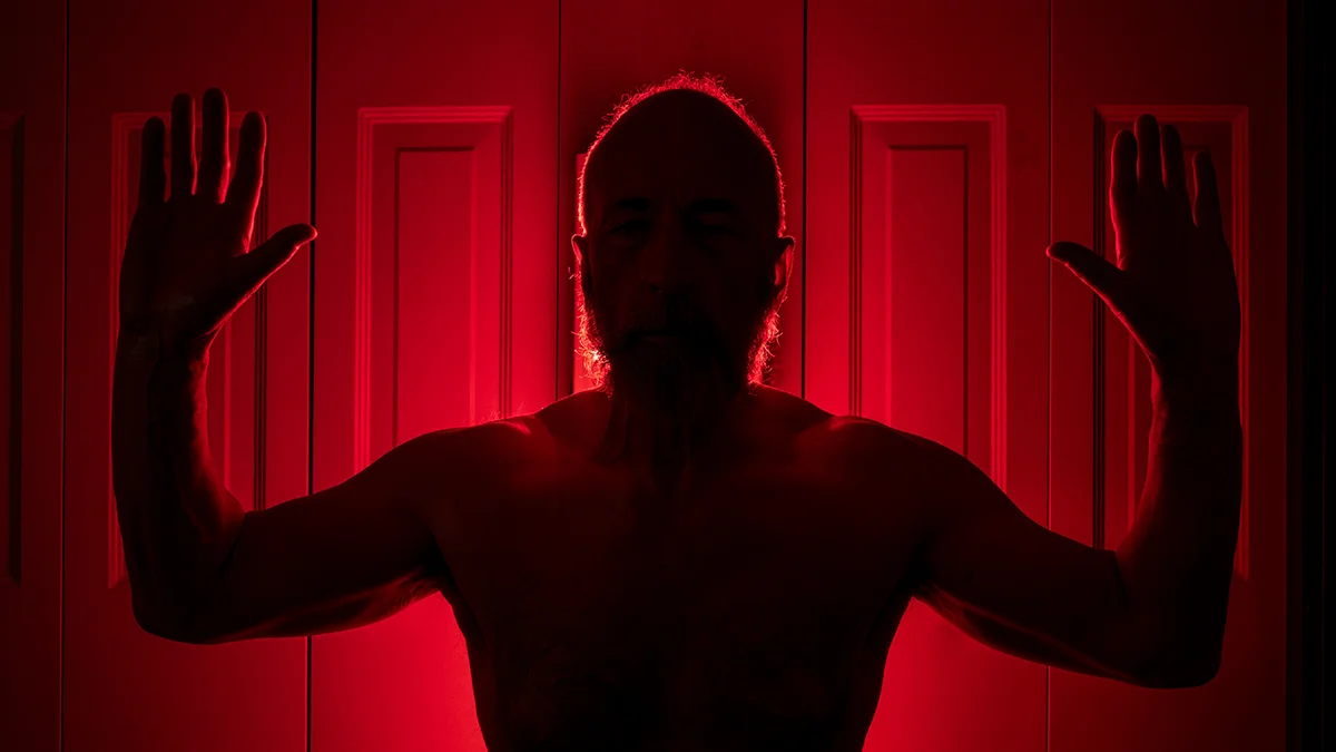 A middle aged man in great shape standing in front of a red light therapy device with arms raised up