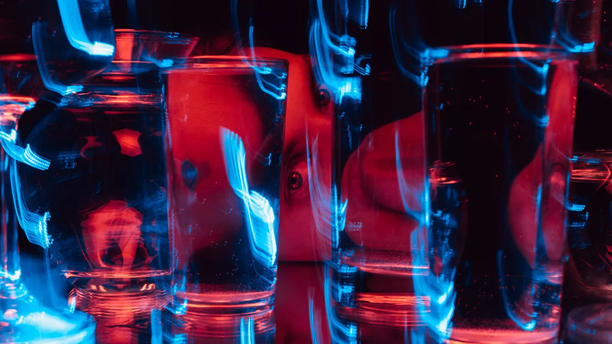 A mans face distorted by glasses of water under red lights on dark background