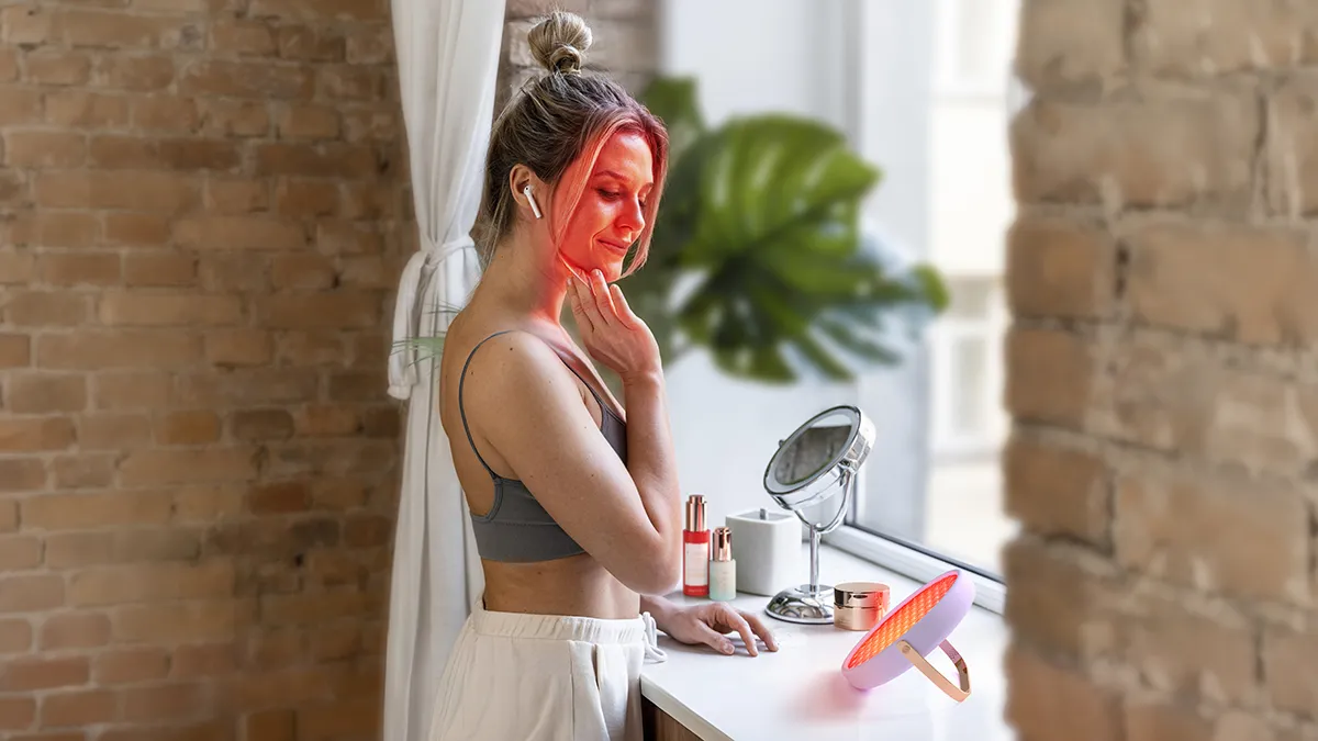 A woman in a tank top standing in front of the window listening to music and using UFO Advanced red light therapy panel