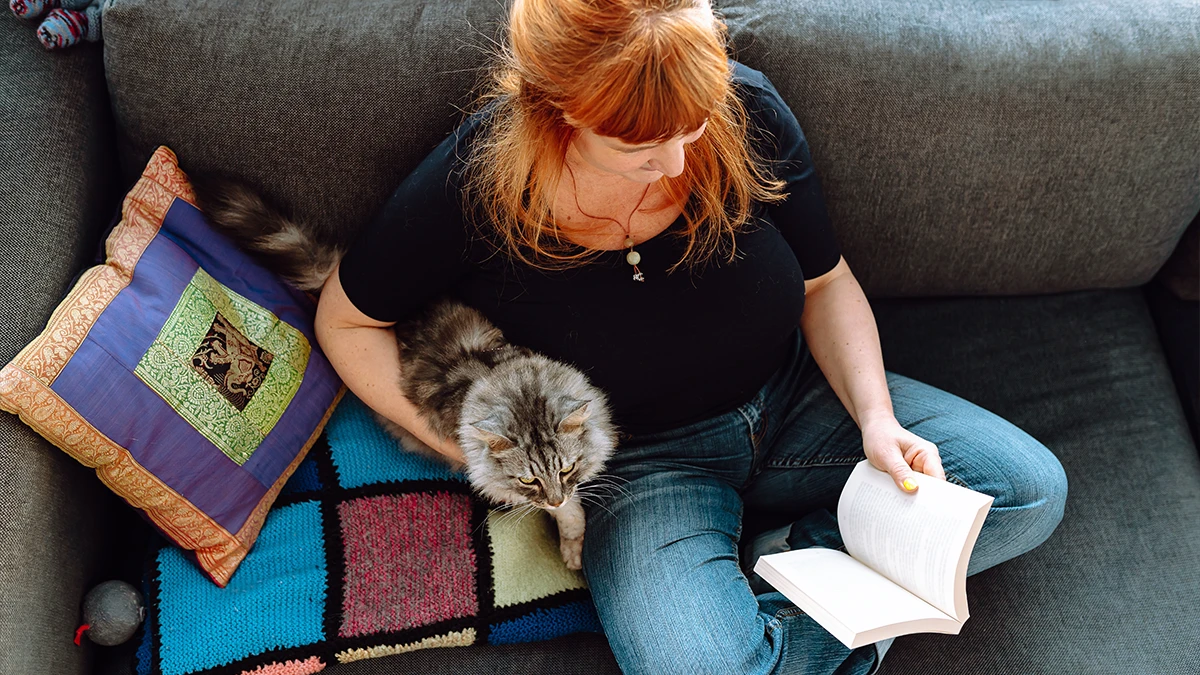 A red headed woman reading a book on her lap and petting a fluffy gray cat