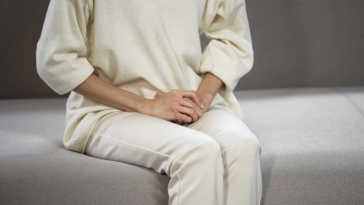 A woman in a white twin set sitting on the edge of the bed, her hands in the lap, covering the vagina