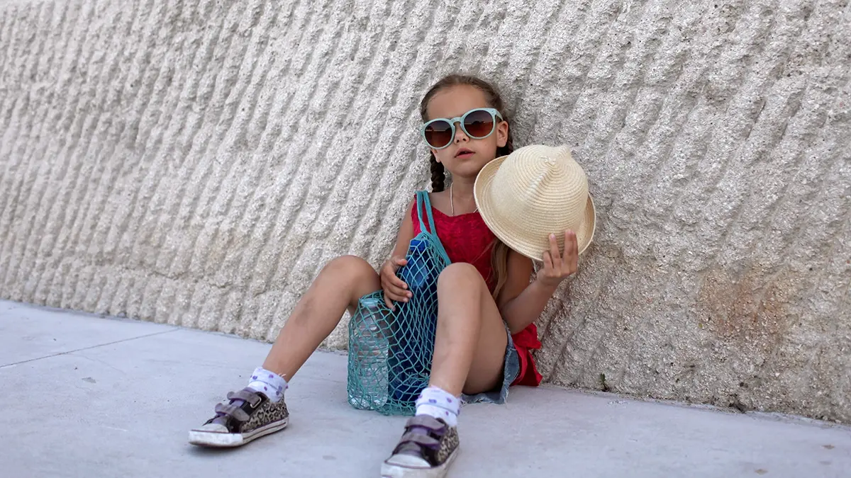 A school girl following advice of how to stay cool in a heatwave witha  hat, sunglases, and a bottle of water, sitting in the shade
