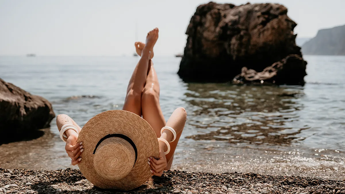 A woman at the beach, on her back and her feet in the air with a large sun hat