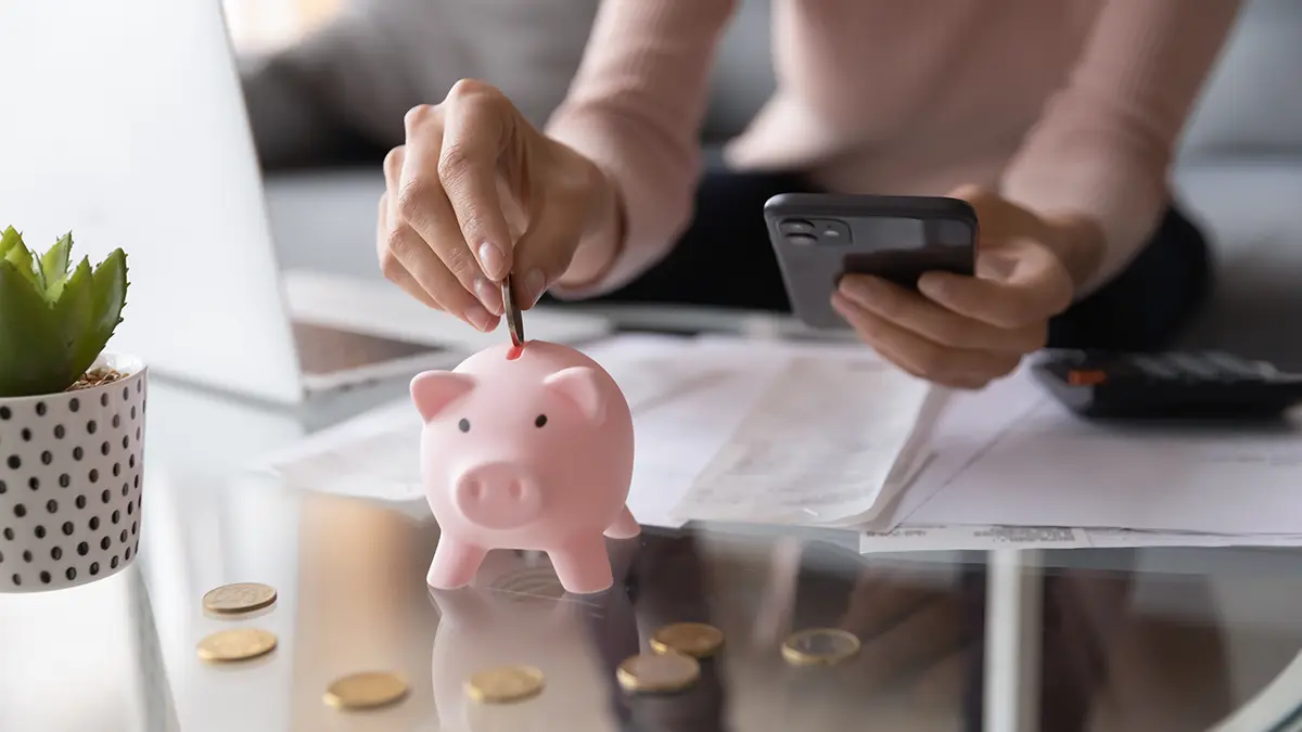 A woman on her phone putting a coin in a piggy bank