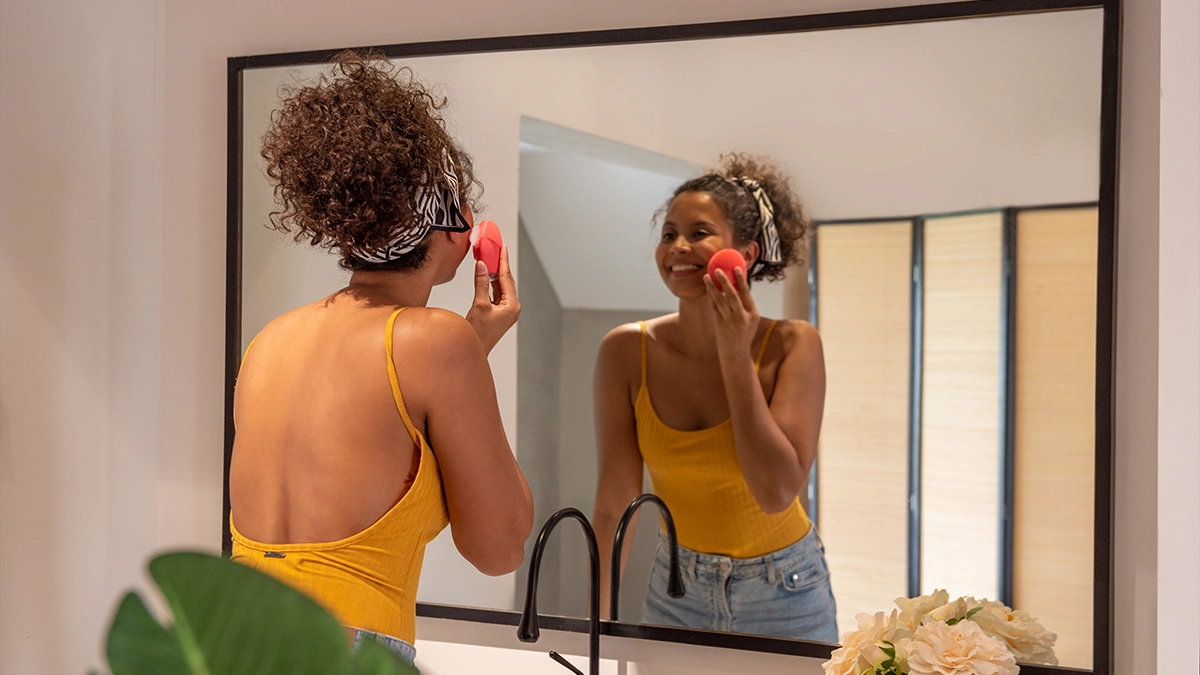 A woman in yellow backless shirt doing a duble cleaning routine with a face brush in the mirror, we see her from the back
