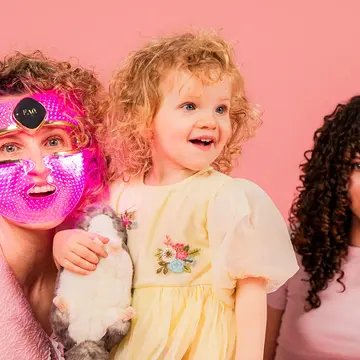 A mom wearing an LED therapy mask with her two daughters, all have curly hair