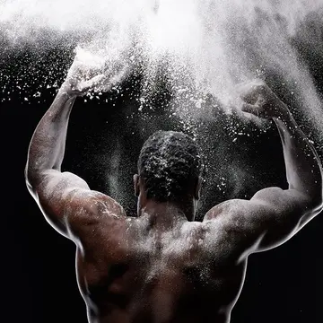 A muscular back of an African-American man clapping his hands covered in white powder in the air on a dark bacground