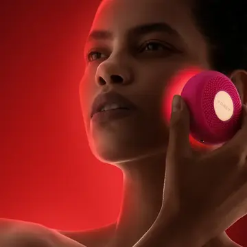 A woman using red LED device therapy on her cheeks on a dark red background