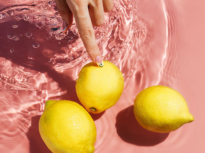 Hand with manicured nails touching one of the three lemons in the water, on a pink surface