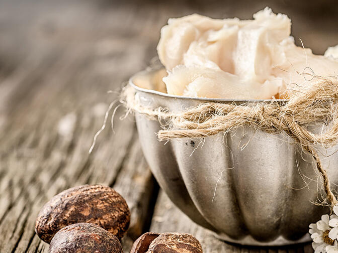 A beautiful textured bowl on an old wooden table, brimming with shea butter, with a shea seed on the left and some wildflowers on the right of the bowl