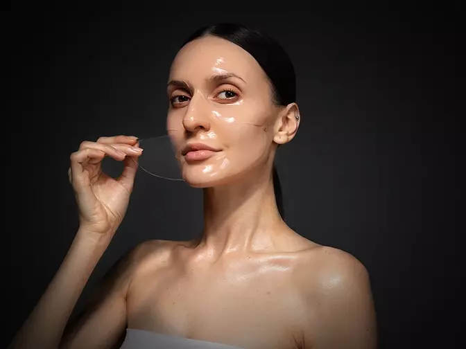 A perfect young woman with sleek dark hair taking the hydrogel mask of her face on dark background A perfect young woman with sleek dark hair taking the hydrogel mask of her face on dark background