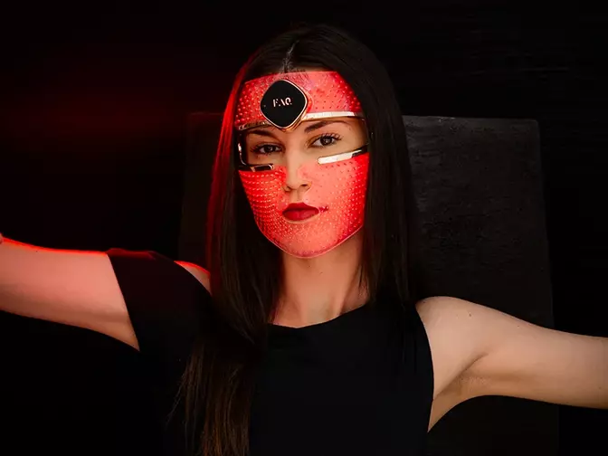 A young woman with dark hair and eyes in a sleveless black dress on black background wearing a red LED face mask 