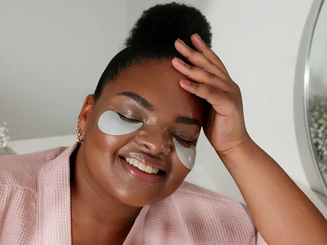 A curvy African-American woman in a pink robe relaxing with hydrating under eye patches