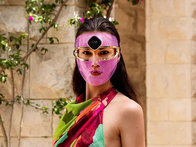 led light therapy what each color does A gorgeous woman with dark hair and dark eyes, in a summer dress, in front of purple flowers, wearing a led face mask set on a purple light
