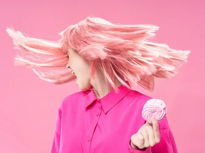 A silver blond-haired woman in pink, on a pink background holding a pink doughnut with hair flying in the air as she turns away from the pastry in her hand