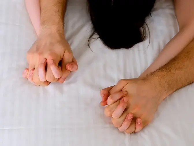 A couple clutching hands during sex on white sheets