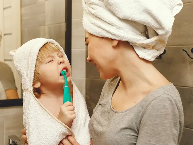 A woman in a hair towel holding a toddler who is brushing his teeth with issa 4 baby toothbrush