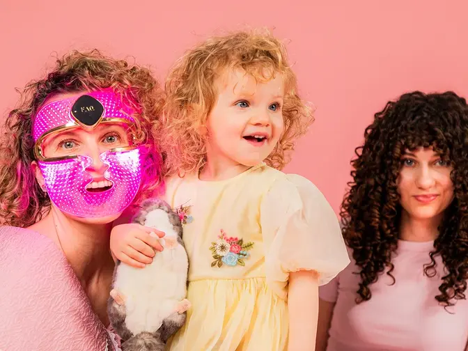 A mom wearing an LED therapy mask with her two daughters, all have curly hair