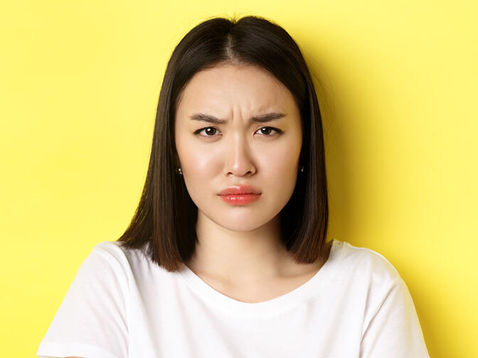 Close up of young Asian woman looking angry and frowning at camera, standing in white t-shirt on yellow background