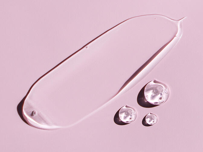 Transparent cosmetic gel or serum smeared over a pink surface alongside three gel drops