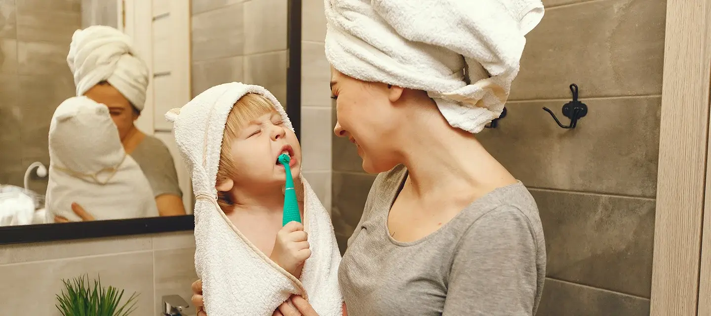 issa 4 baby A woman in a hair towel holding a toddler who is brushing his teeth with issa 4 baby toothbrush