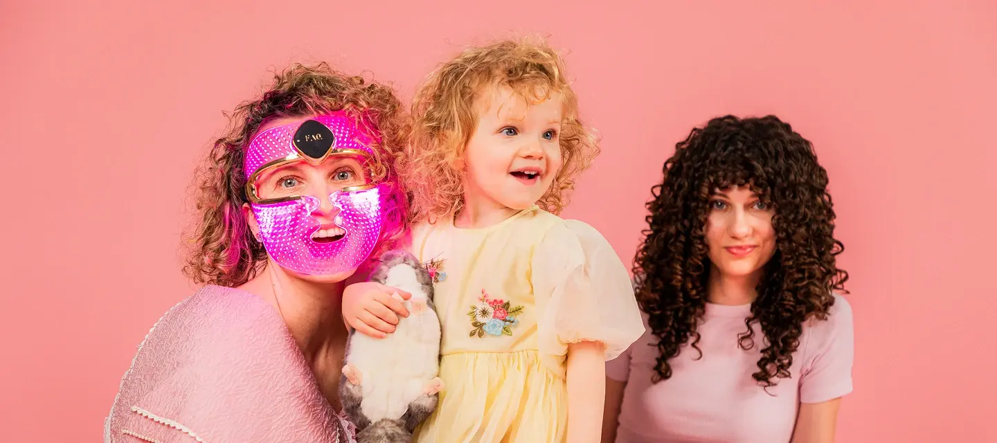 A mom wearing an LED therapy mask with her two daughters, all have curly hair