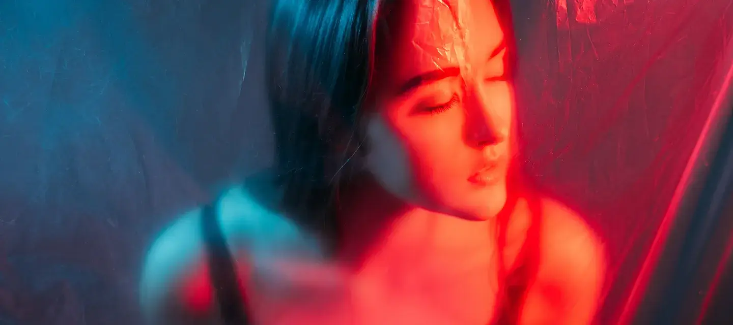 A woman behind a translucent barrier, eyes closed and illuminated by red light therapy