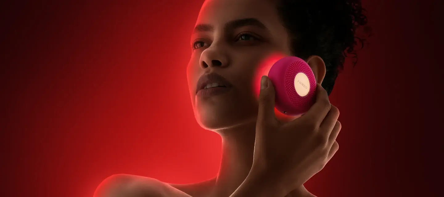 A woman using red LED device therapy on her cheeks on a dark red background