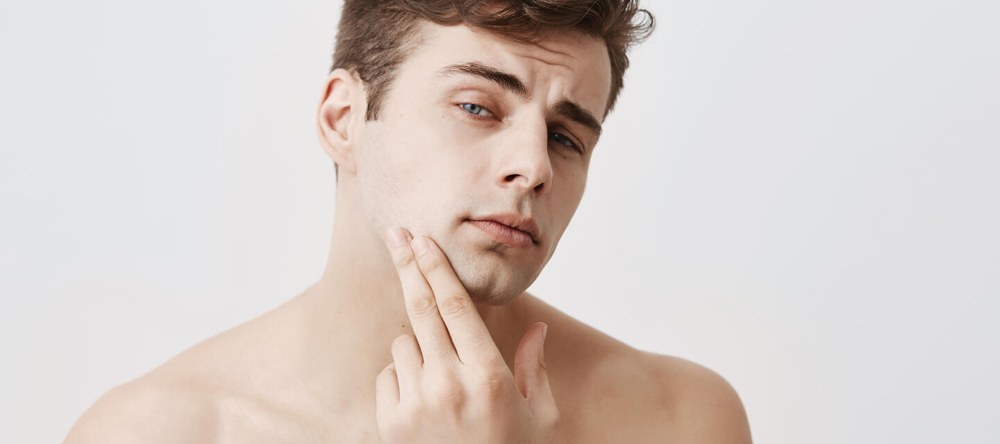 Handsome attractive young man with dark hair, stylish haircut, blue eyes, keeps hand on chin, looks pensively at camera, isolated against gray background. Confident naked european guy with pure and healthy skin Handsome attractive young man with dark hair, stylish haircut, blue eyes, keeps hand on chin, looks pensively at camera, isolated against gray background. Confident naked european guy with pure and healthy skin
