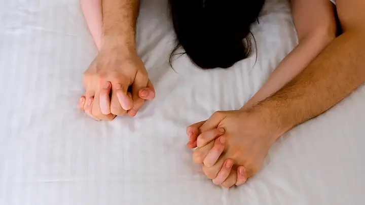 A couple clutching hands during sex on white sheets