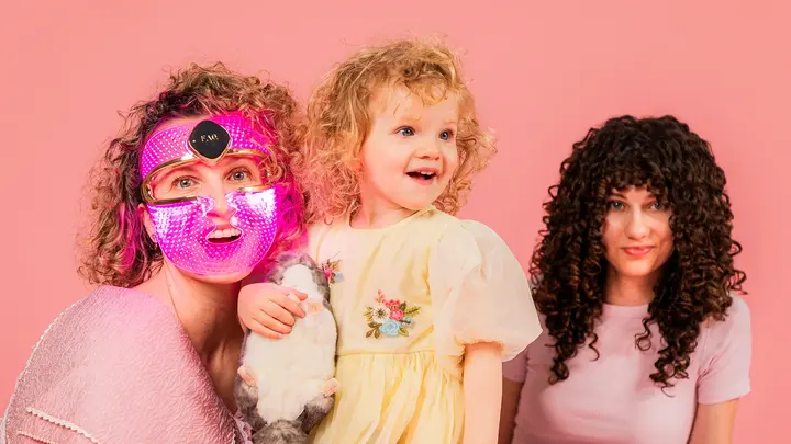 A mom wearing an LED therapy mask with her two daughters, all have curly hair