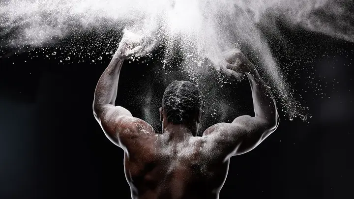A muscular back of an African-American man clapping his hands covered in white powder in the air on a dark bacground