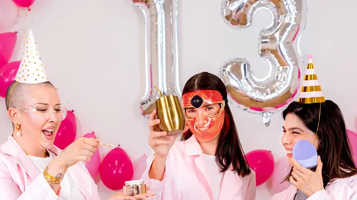 Three young women celebrating FOREO birthday sale with drinks and doing skincare in a white and pink setting
