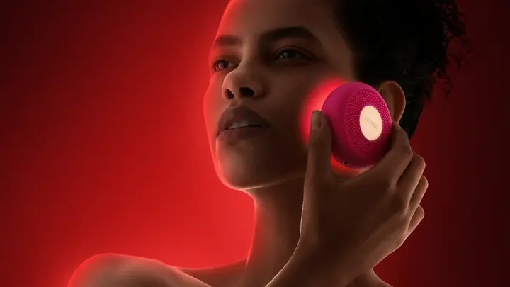 A woman using red LED device therapy on her cheeks on a dark red background