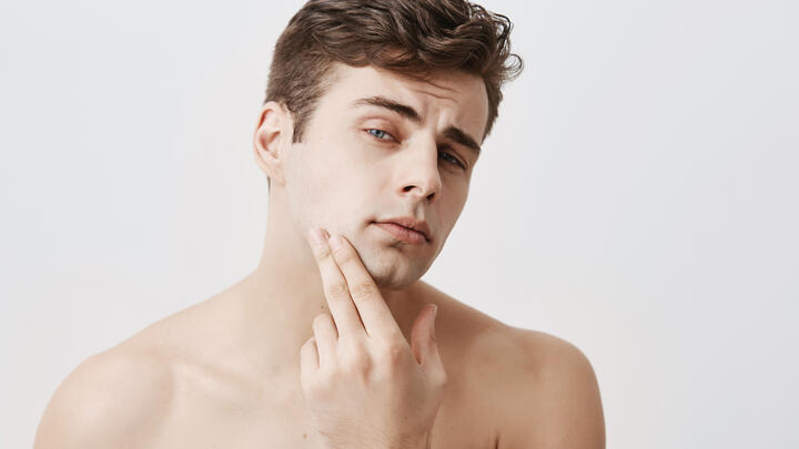 Handsome attractive young man with dark hair, stylish haircut, blue eyes, keeps hand on chin, looks pensively at camera, isolated against gray background. Confident naked european guy with pure and healthy skin